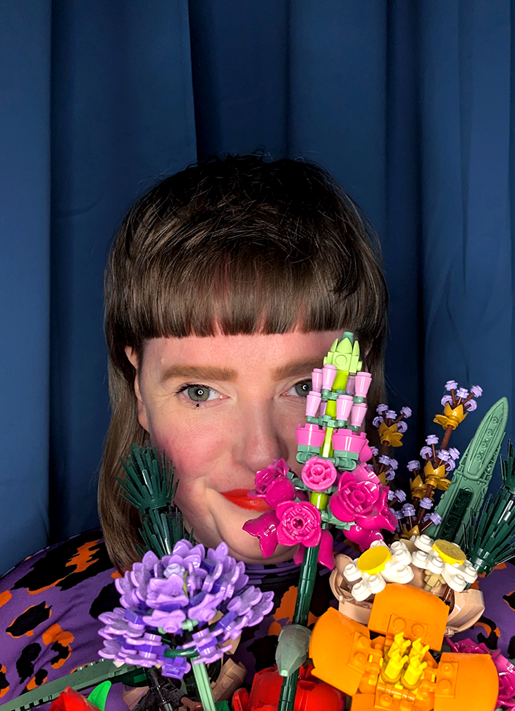 Mii hält einen bunten LEGO-Blumenstrauß vor ihr Gesicht, sodass nur ihre Augen und ein Teil ihres Gesichts sichtbar sind. Sie trägt einen lilafarbenen Leopardenmuster-Pullover auf orangem Grund. Der Hintergrund ist ein tiefblauer Vorhang.